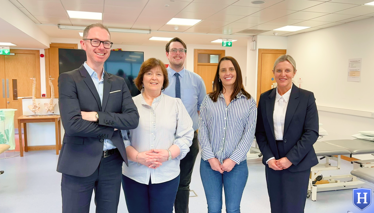 A group of five professionals from HealthCo (Left to right: Declan Savage, Susan Manning, Patrik Taliga, Aishling Roche, Majella Savage) standing in a medical training room, smiling at the camera. The team, dressed in business and smart casual attire, provided Basic Life Support (BLS) and Patient Handling Training, equipping students with essential life-saving and patient mobility skills.